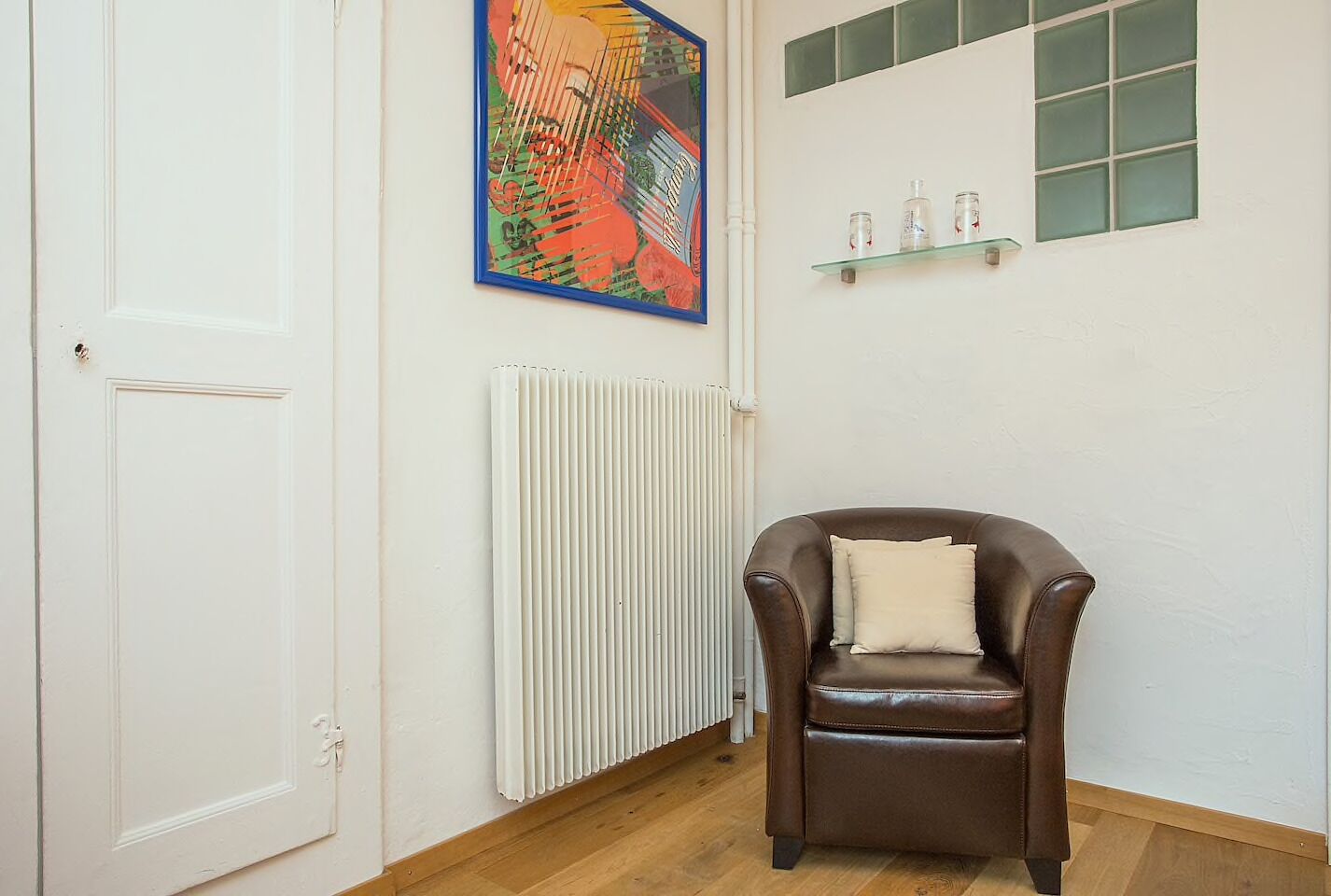 A room with a brown leather chair, a radiator, a white door, a colorful painting, and a shelf with bottles.