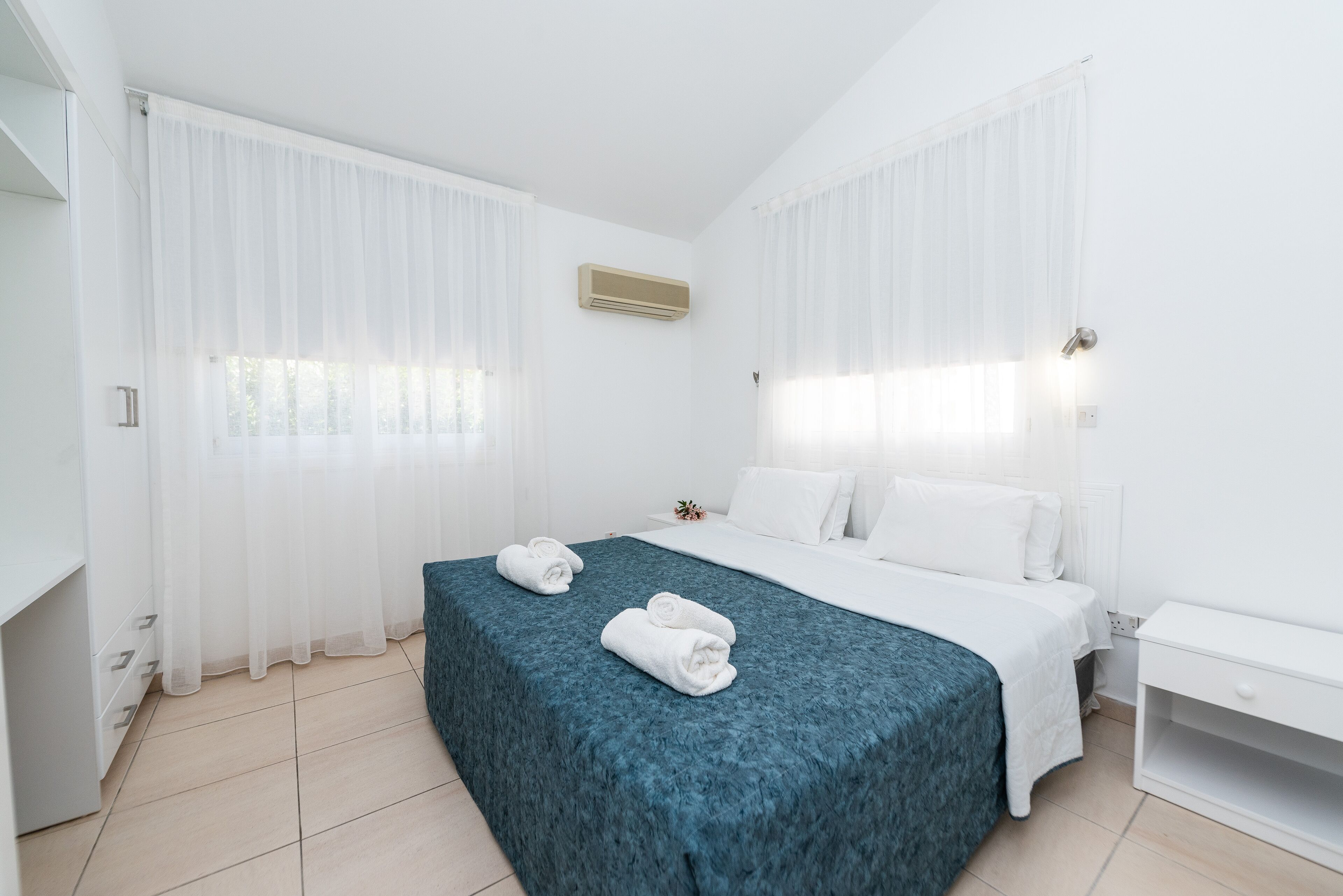 Et hotelværelse med seng, et natbord, et vindue med let gardin og en aircondition.