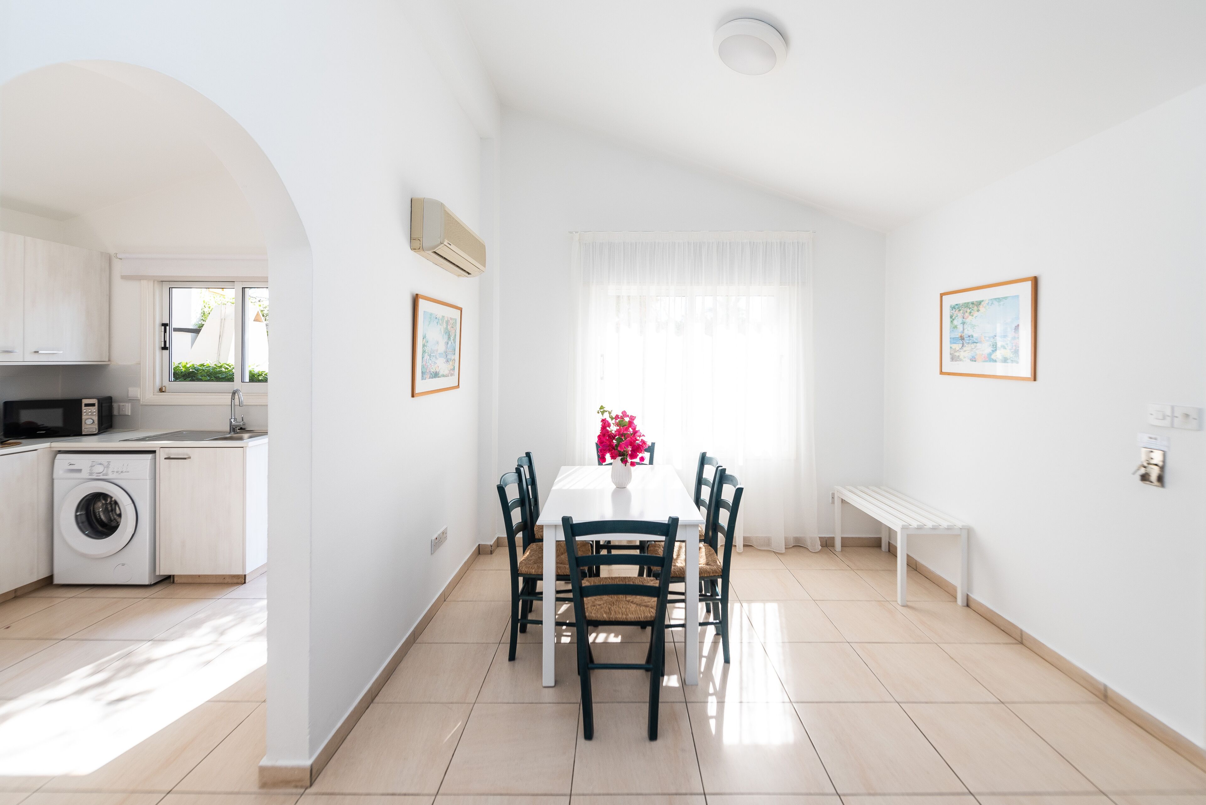 A bright, white room with a dining table, chairs, and a bench. There is a washing machine and a window with curtains.