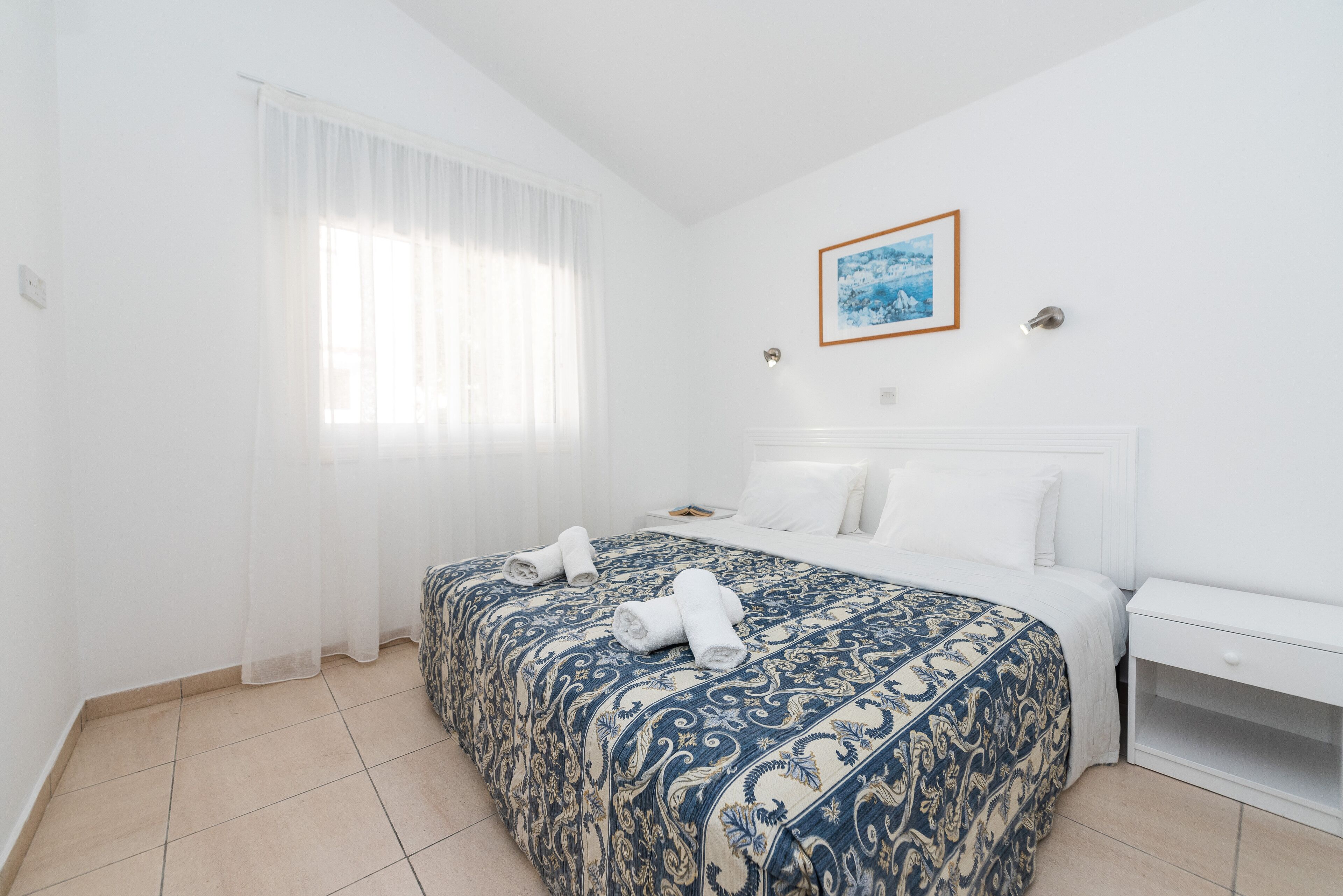 Quarto de hotel com uma cama grande, uma mesa de cabeceira, um aparador branco e uma janela com cortinas leves.