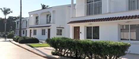 Una hilera de edificios blancos de dos pisos con balcones, una acera pavimentada y palmeras.