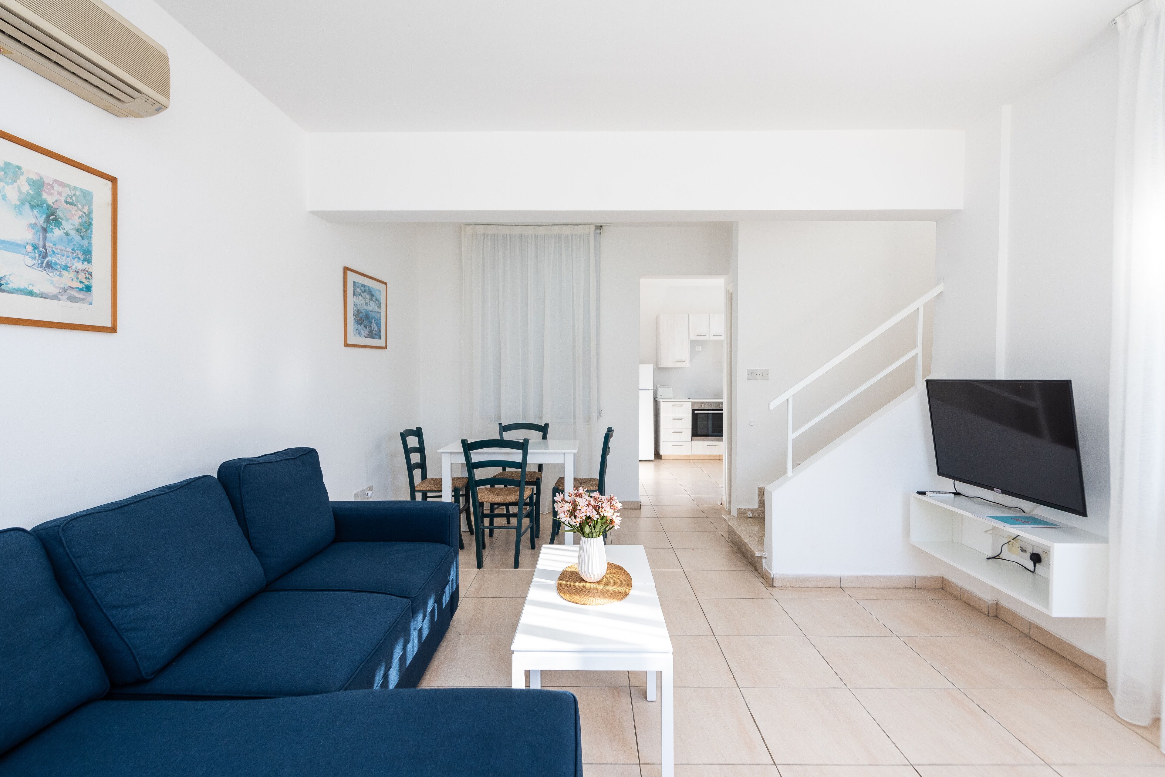 A modern living room with a blue sofa, a white coffee table with a vase, a flat-screen TV, and a staircase leading to a kitchen.