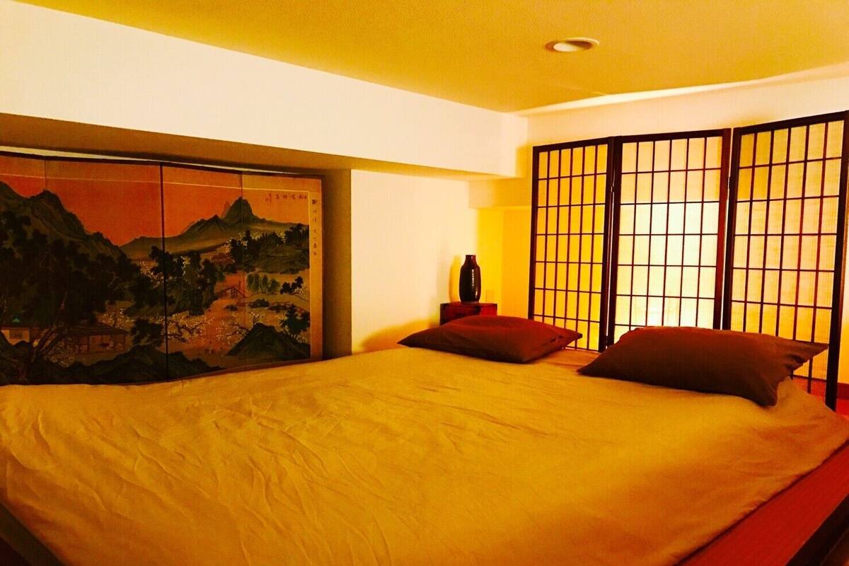 A bedroom with a large bed, a painting on the wall, and a traditional Japanese shoji screen.