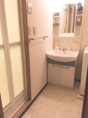 A small bathroom with a toilet, sink, and mirror. There is a towel rack above the sink and a small shelf with toiletries. - Local Nakasu Apartment (Fukuoka)