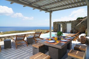Un área de comedor al aire libre con vista al mar, paredes de piedra y una pérgola.