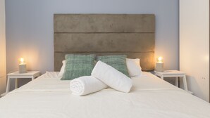 Una cama bien tendida con sábanas blancas, dos almohadas verdes y una toalla blanca doblada. Hay dos lámparas de noche en mesitas blancas.