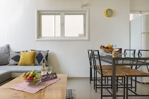 A modern living room with a sofa, a dining table with chairs, a window, and a clock on the wall. - Sweet Home At Hilton Beach 2BR (Tel Aviv)