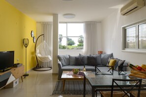 A modern living room with a sofa, a glass coffee table, a TV, and a hanging chair. - Sweet Home At Hilton Beach 2BR (Tel Aviv)