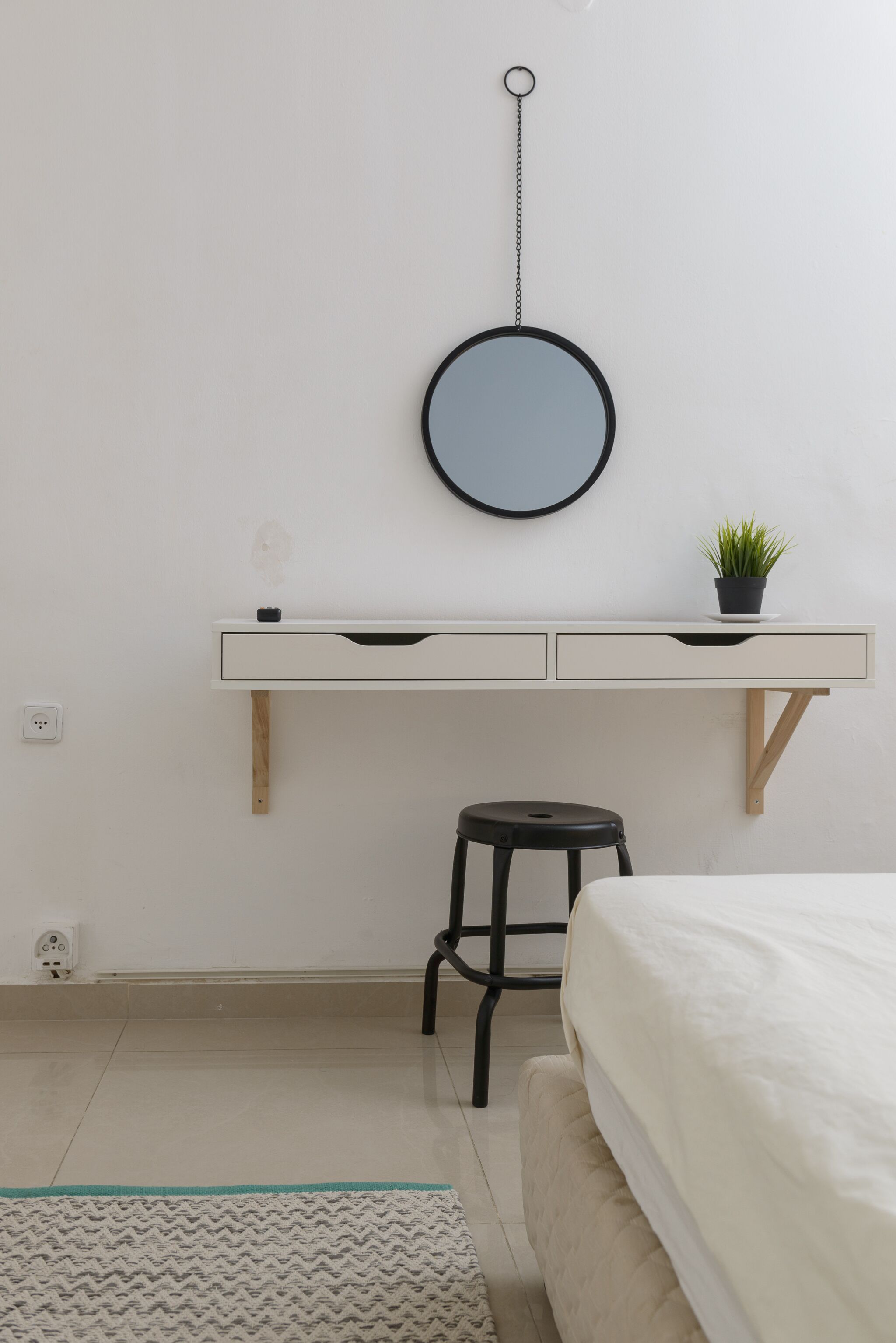 Een minimalistische slaapkamer met een bed, een bureau met spiegel, een klein krukje en een potplant.