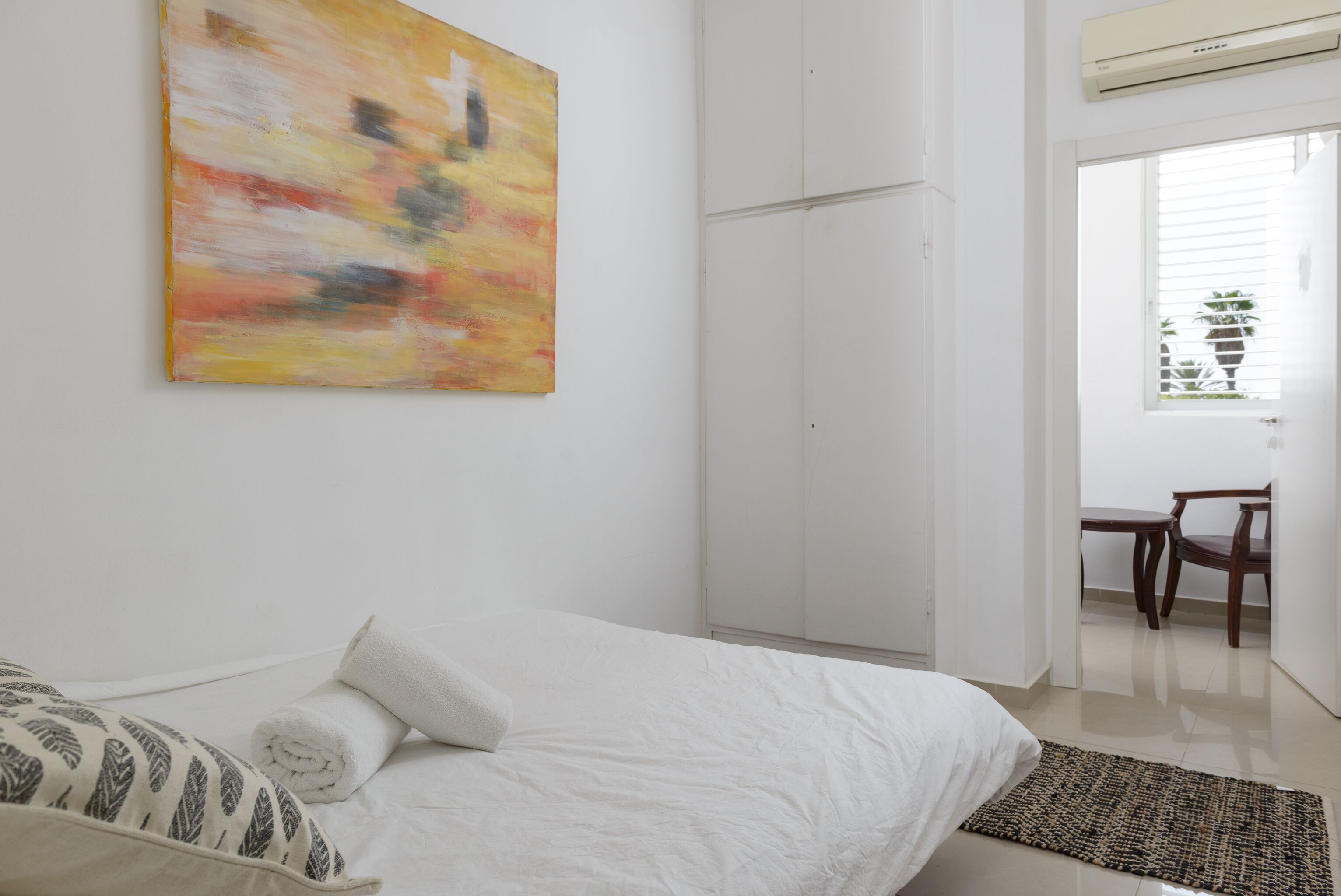 A bedroom with a bed, a white wall, a painting, and a door leading to a dining area.