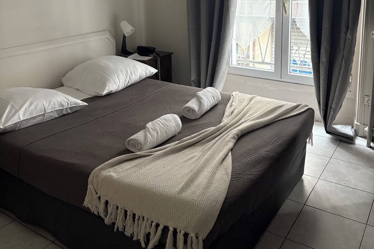 A neatly made bed with white pillows and a beige blanket, a bedside table with a lamp, and a view of the outside through large windows with grey curtains.