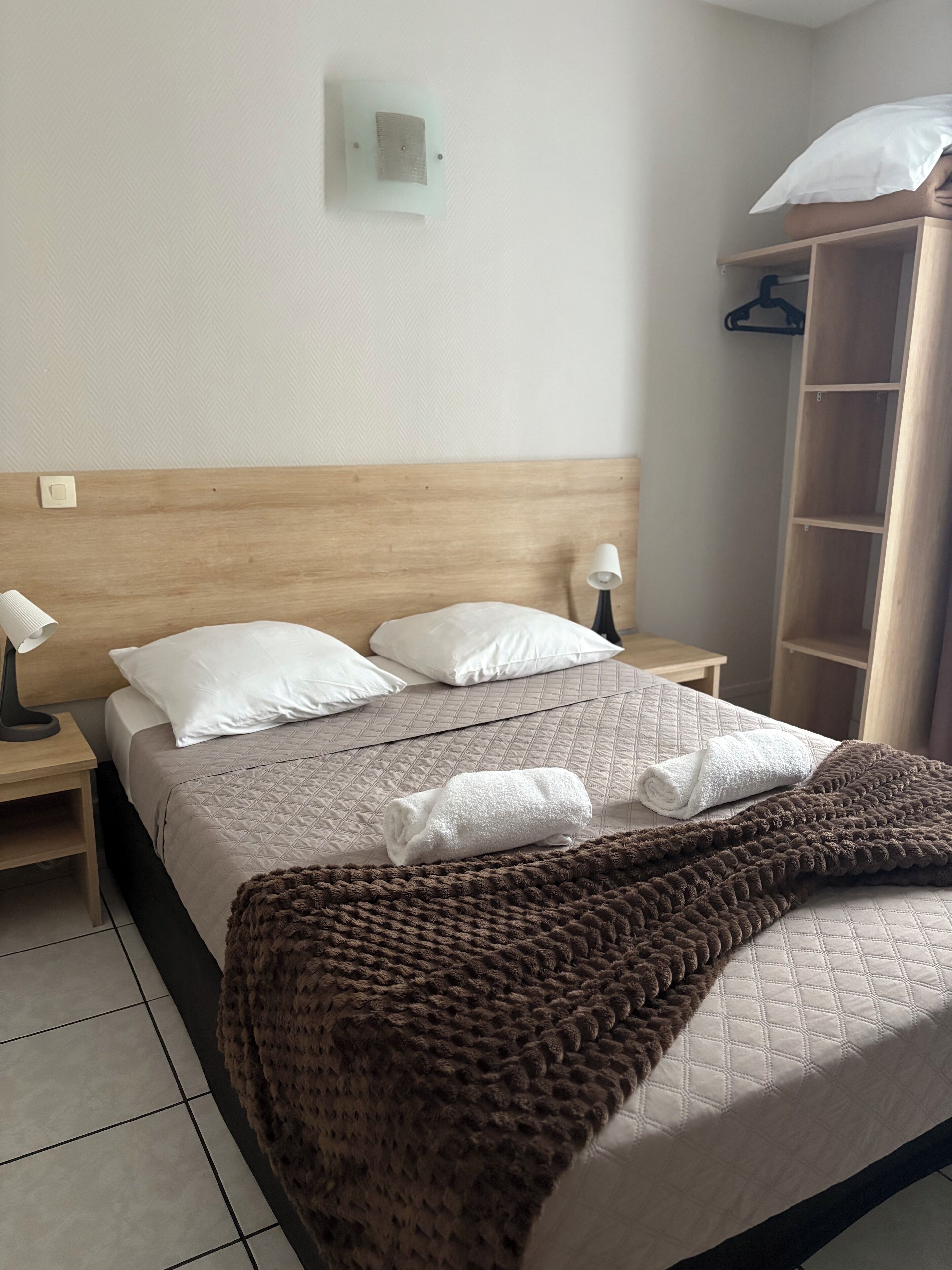 A neatly made bed with a brown blanket, white pillows, and a quilted headboard. There are two bedside tables with lamps and a wall-mounted light switch.