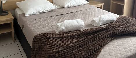 A neatly made bed with a brown blanket, white pillows, and a quilted headboard. There are two bedside tables with lamps and a wall-mounted light switch.