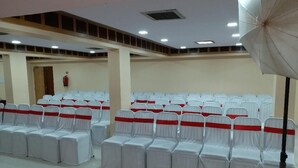 A conference room with rows of white chairs with red sashes, a ceiling with recessed lighting, and a fire extinguisher on the wall. - Hotel Three Star Pvt Ltd (Panvel)