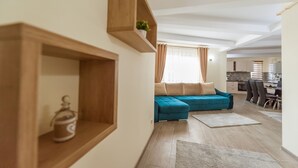 A modern living room with a teal sofa, wooden shelves, and a kitchen in the background.
