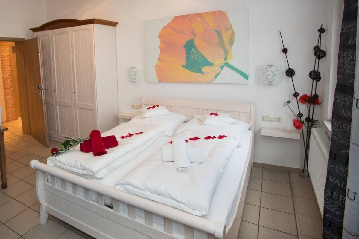 Una cama bien tendida con sábanas blancas y pétalos de rosa roja, un armario blanco y un cuadro de una flor en la pared.