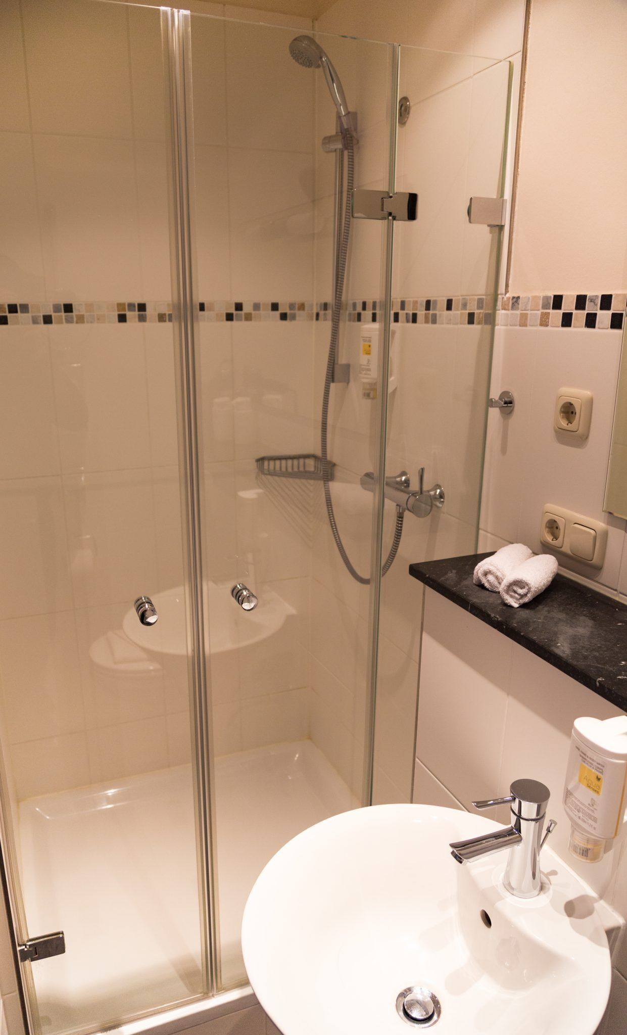 A glass-enclosed shower with a handheld showerhead, a glass door, and a tiled wall with a mosaic border.