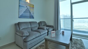 Un salon moderne avec un canapé gris, une table basse en bois, un panneau d’accueil et une grande fenêtre donnant sur des immeubles.
