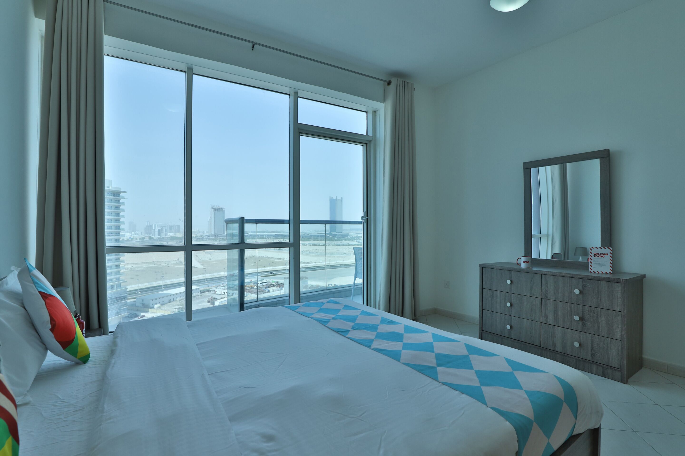 Une chambre d’hôtel avec une grande fenêtre, un lit recouvert d’une courtepointe à carreaux, une commode et un miroir.