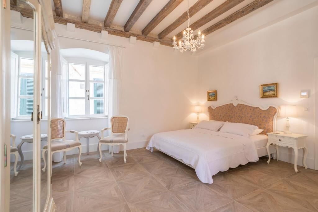 Un dormitorio con una cama grande, dos mesitas de noche, una araña y vistas al exterior a través de una ventana con contraventanas.