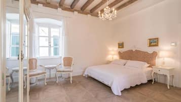 Un dormitorio con una cama grande, dos mesitas de noche, una araña y vistas al exterior a través de una ventana con contraventanas.