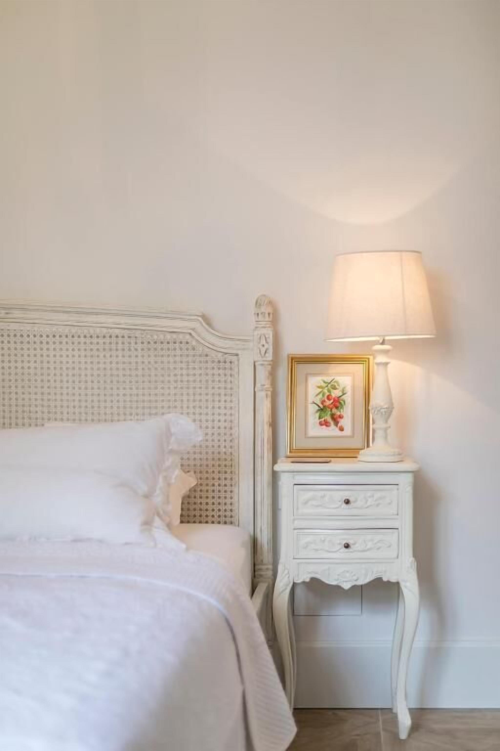 Un lit bien fait, avec du linge de lit blanc, une table de chevet blanche ornée d’une lampe et d’un tableau encadré, et une tête de lit classique en rotin.