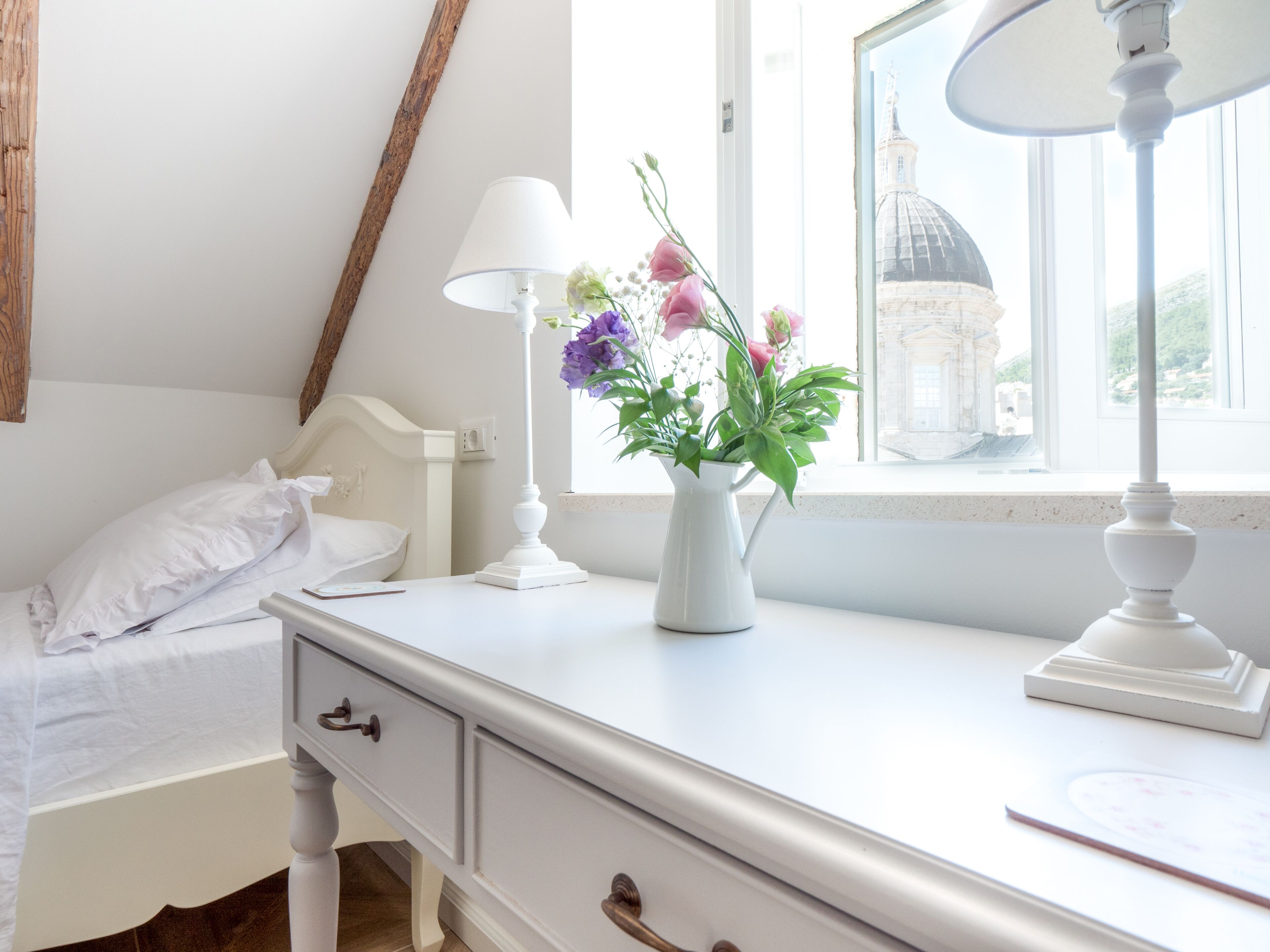Een slaapkamer met een bed, nachtkastje, lamp, vaas met bloemen en een raam met uitzicht op een gebouw.