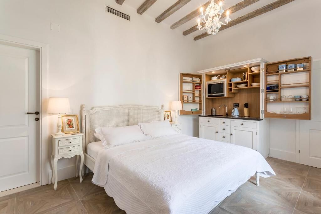 Een slaapkamer met een wit bed, nachtkastjes met lampen, een inbouwkast voor de televisie en een houten kast met servies.