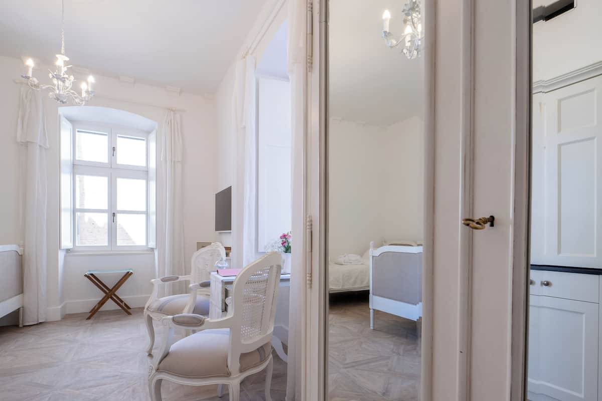 Quarto com mesa de jantar, cadeiras brancas, uma cama e janela com cortinas.