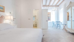 Een slaapkamer met een groot bed, een houten vloer, een zithoek met stoelen en een keuken zichtbaar door een open deur.