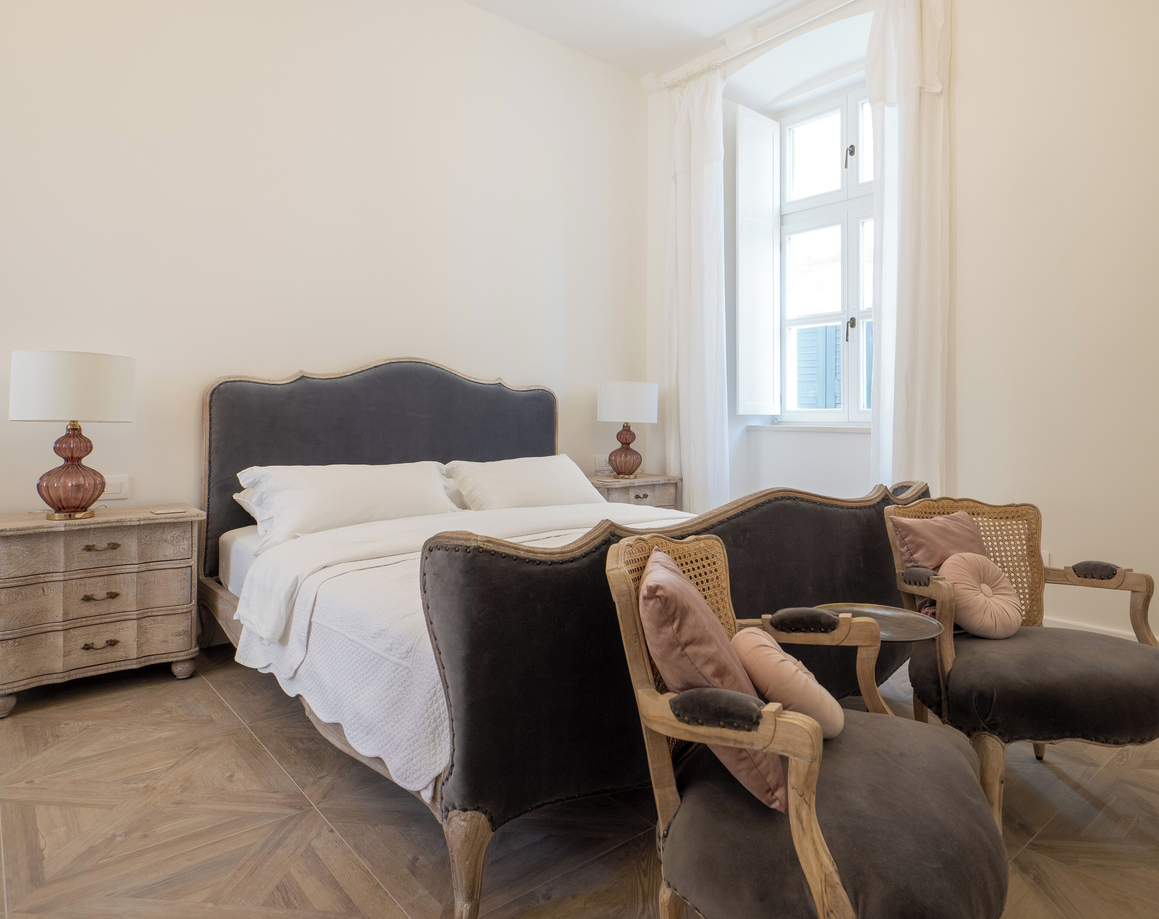 Quarto com cama, duas mesas de cabeceira, janela e duas poltronas estofadas.