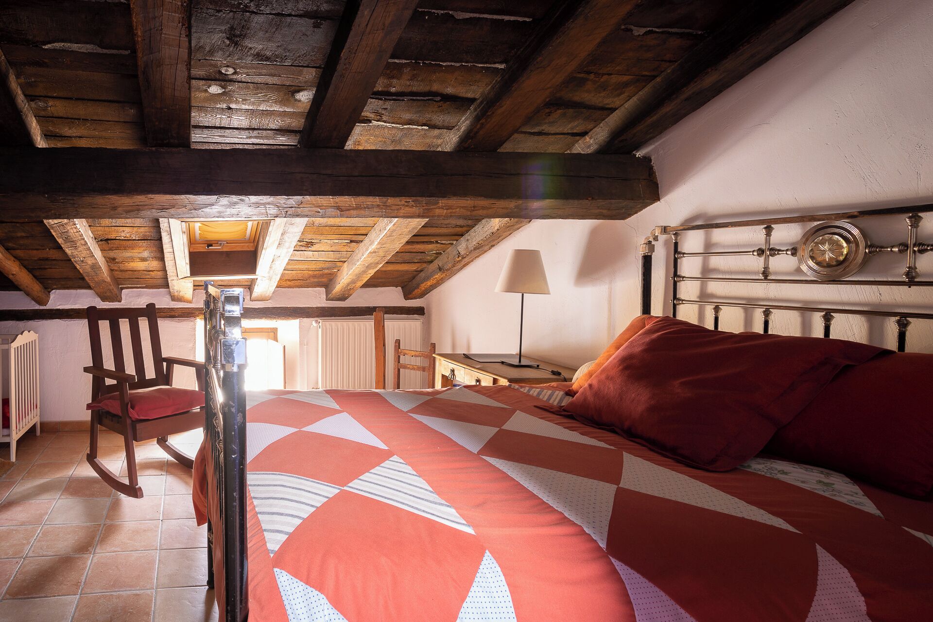 A bedroom with a wooden ceiling, a bed with a patterned bedspread, a chair, a bedside table with a lamp, and a wall clock.