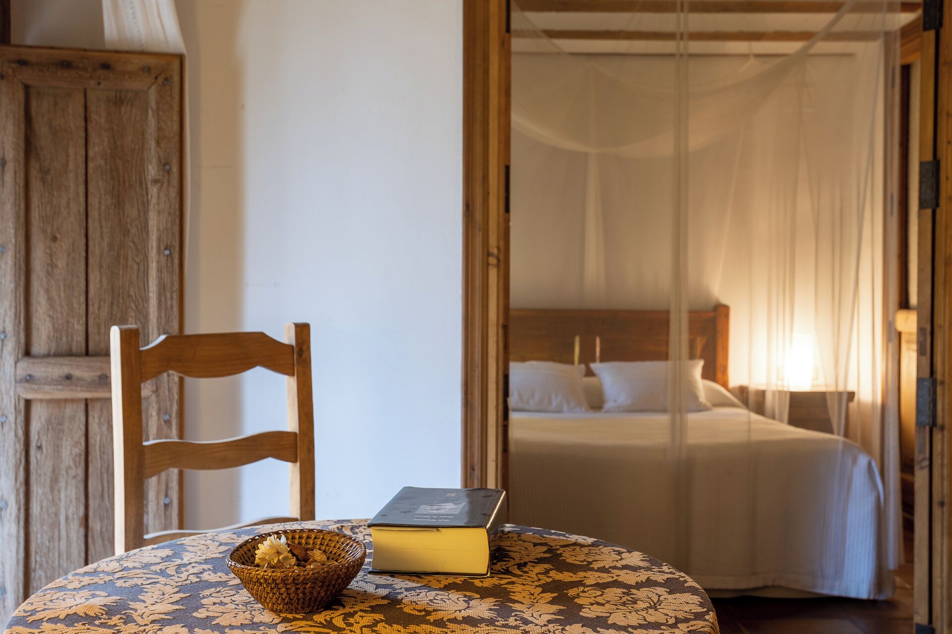 A room with a bed, a chair, a table with a book and a basket.