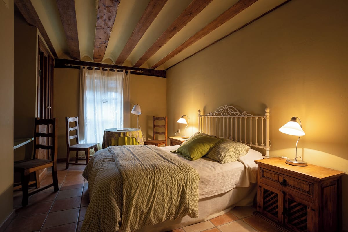 Een slaapkamer met een bed, twee nachtkastjes met lampen, een stoel, een klein tafeltje en een raam met gordijnen.
