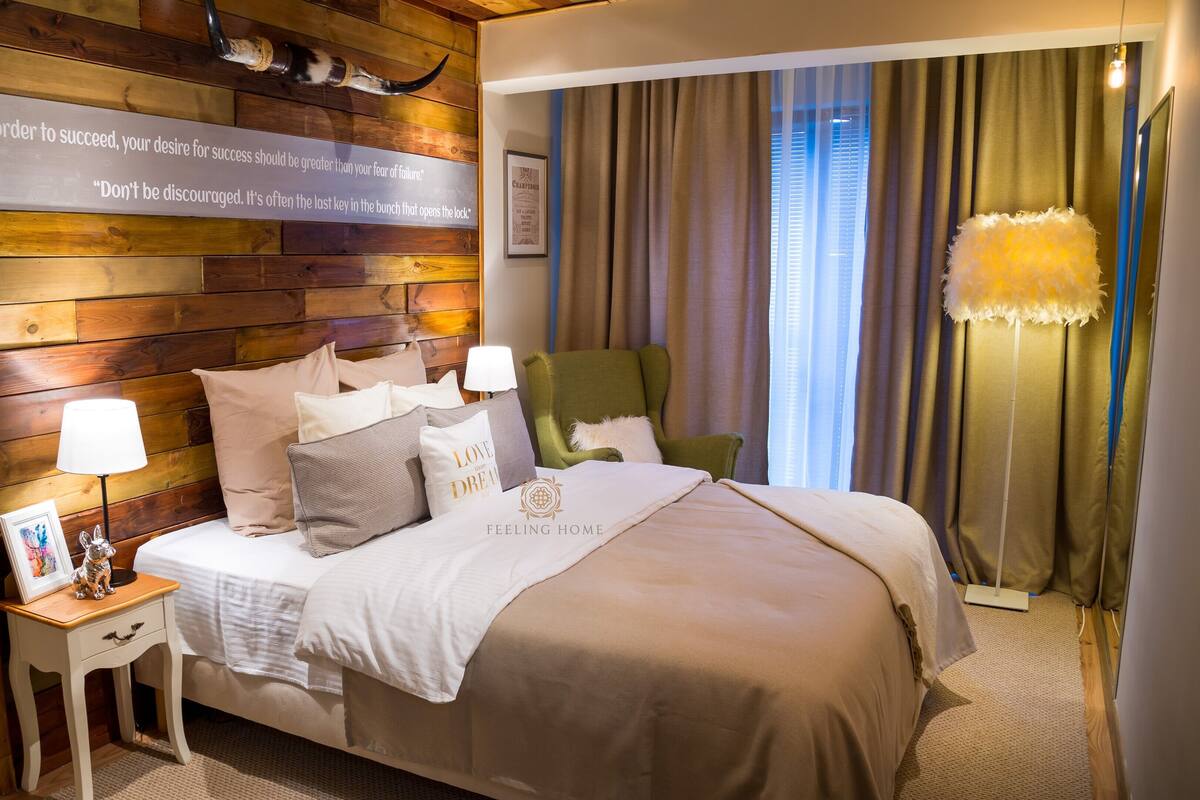 Ein Hotelzimmer mit einem Holzkopfteil, einem Bett mit vielen Kissen, einem Nachttisch mit Lampe, einem grünen Sessel und einem Fenster mit Vorhängen.