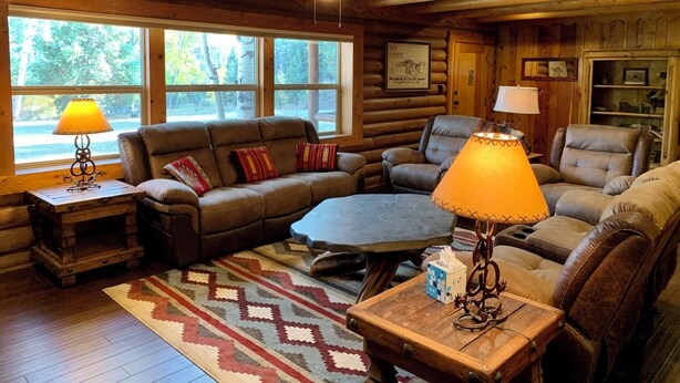 L’intérieur d’un chalet confortable, avec des murs en bois, une grande fenêtre, une cheminée et un coin salon accueillant, comprenant une table basse et des lampes.