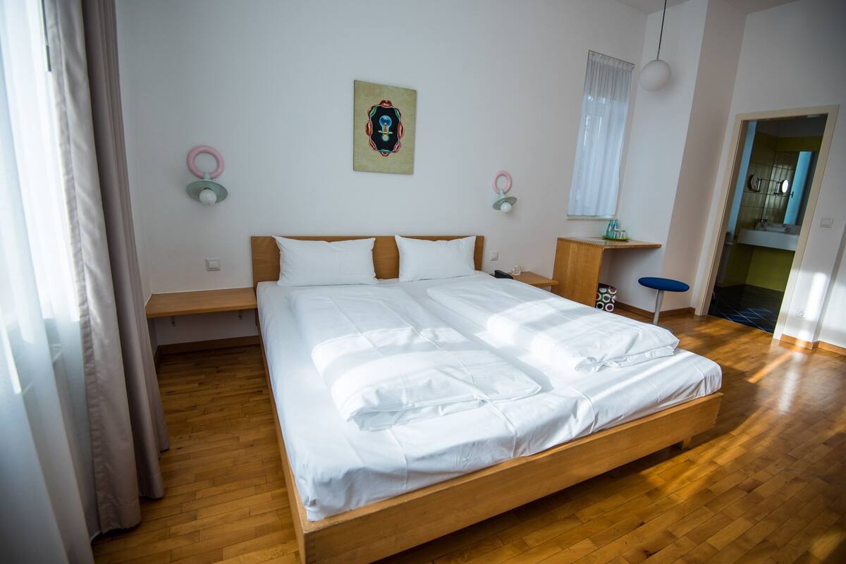 Ein Schlafzimmer mit einem hölzernen Bett, weißer Bettwäsche, einem Nachttisch, einer Wandleuchte und einem Vorhangfenster.
