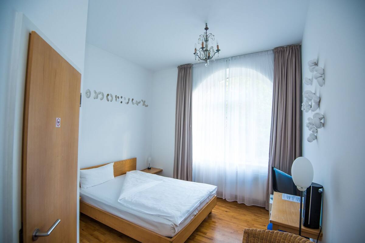 Ein Schlafzimmer mit einem hölzernen Bett, einem Schreibtisch mit Computer, einem Sessel, einer Chandelierleuchtre und einem Fenster mit Vorhängen.