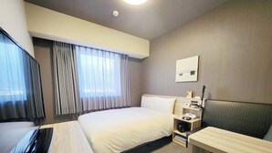 Una camera d'albergo con un letto, una scrivania, una sedia, una TV, un comodino con una lampada e una finestra con tende.