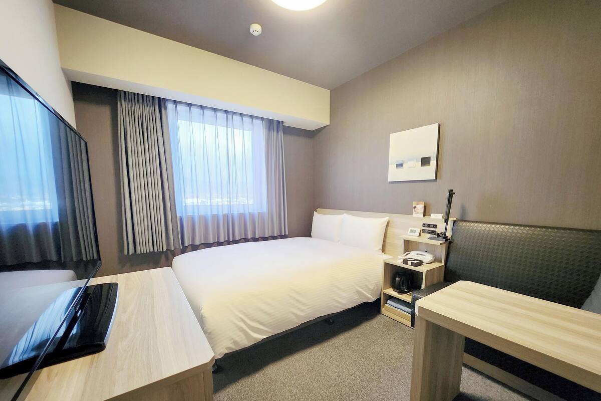 A hotel room with a bed, a desk, a chair, a TV, a bedside table with a lamp, and a window with curtains.