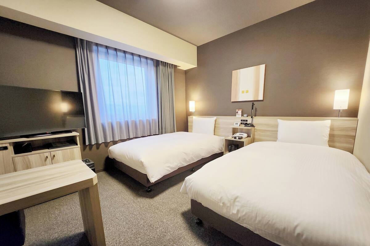 A hotel room with two beds, a TV, a desk, and a nightstand with a lamp.