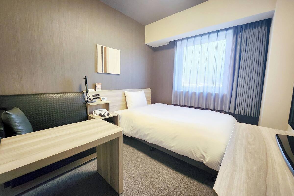 A hotel room with a bed, a desk, a chair, a nightstand, and a window with curtains.