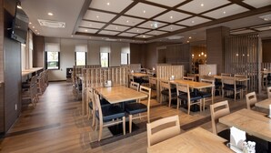 Un ristorante moderno con tavoli e sedie in legno, un soffitto alto con illuminazione a incasso e vista sull'esterno attraverso ampie finestre.
