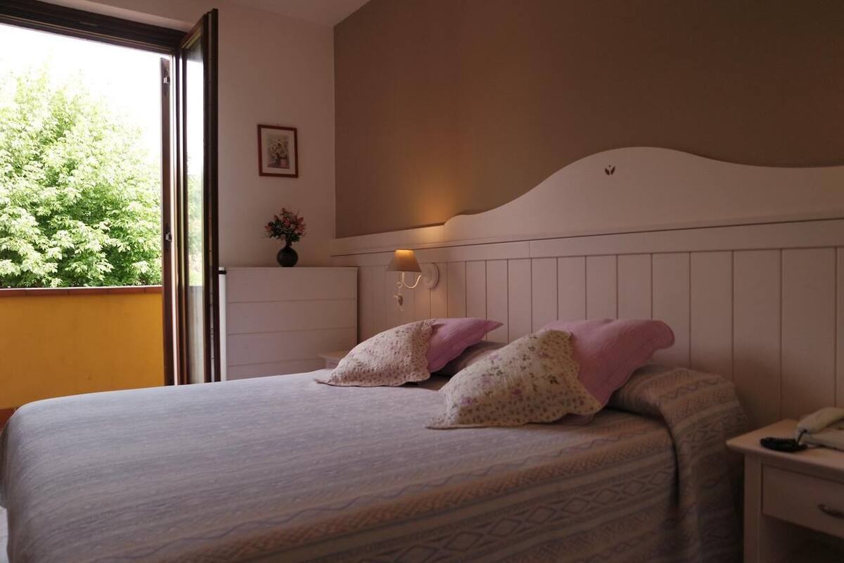 A bedroom with a bed, pillows, a bedside table with a lamp, a wall-mounted light, and a window with a view of trees.
