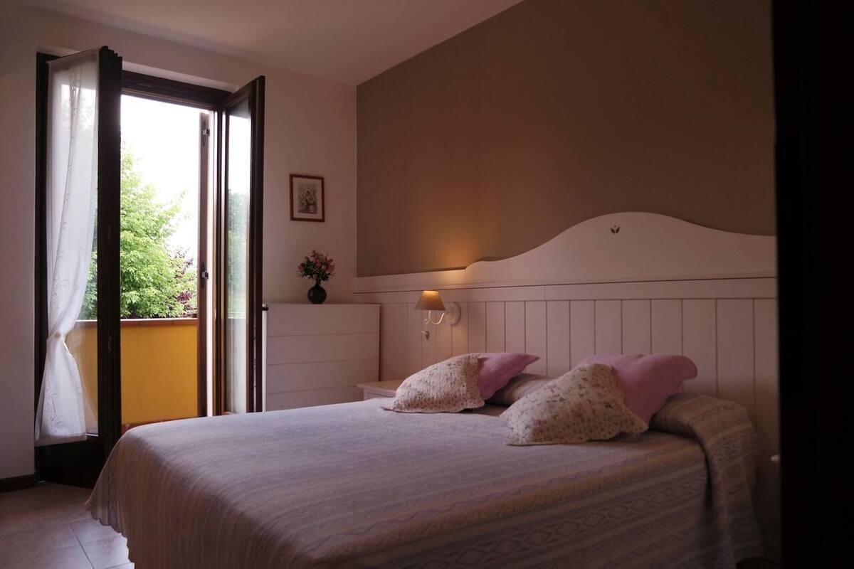A bedroom with a bed, pillows, a nightstand, a wall-mounted lamp, and a window with curtains.
