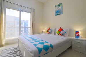 Una habitación de hotel con cama, mesita de noche, ventana con cortinas y un cuadro en la pared, con vistas a edificios.