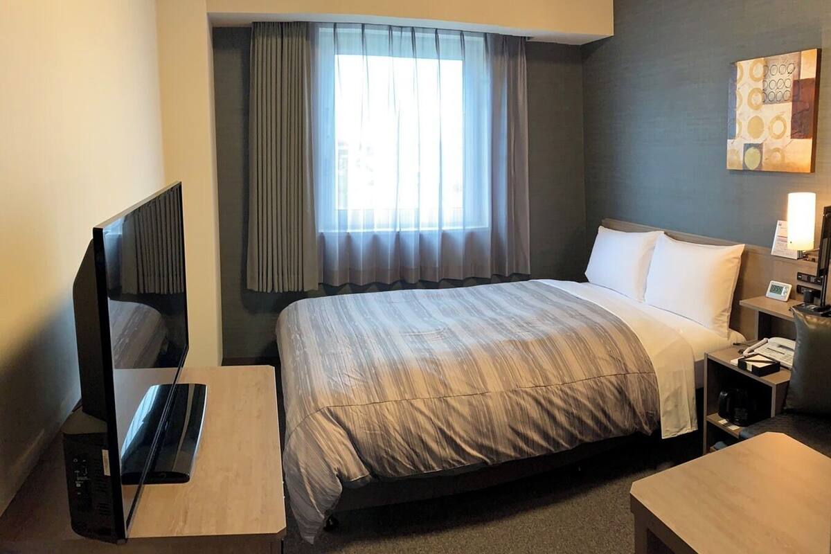 A hotel room with a bed, a desk with a computer, a TV, a bedside table with a lamp, and a window with curtains.