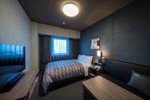 A hotel room with a large bed, a desk, a TV, a bedside table with a lamp, and a window with curtains.