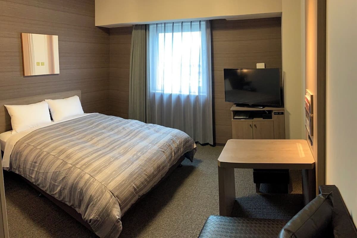 A hotel room with a large bed, a TV, a small table, and a window with curtains.