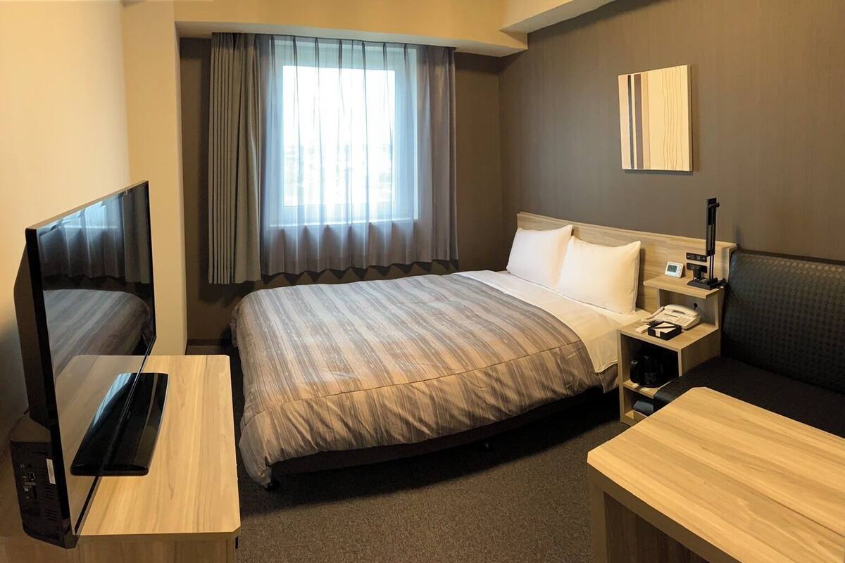 A hotel room with a bed, a TV, a desk, a bedside table, a lamp, and a window with curtains.